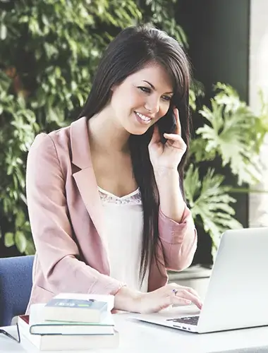 Smiling woman working on a laptop while talking on the phone in a modern office.